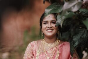 A smiling South Indian bride in a pink saree adorned with gold jewelry, captured outdoors.